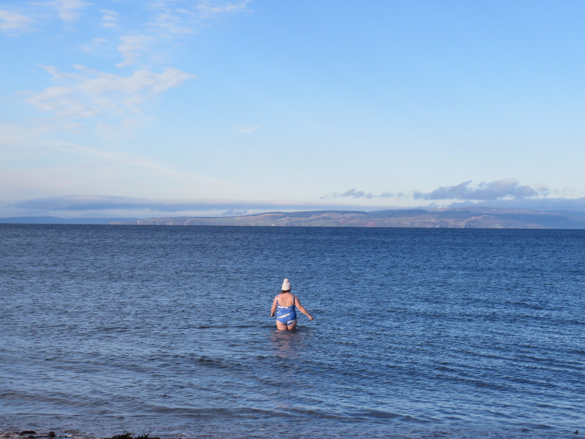 A person in a bathing suit and woolly hat walks out into the sea