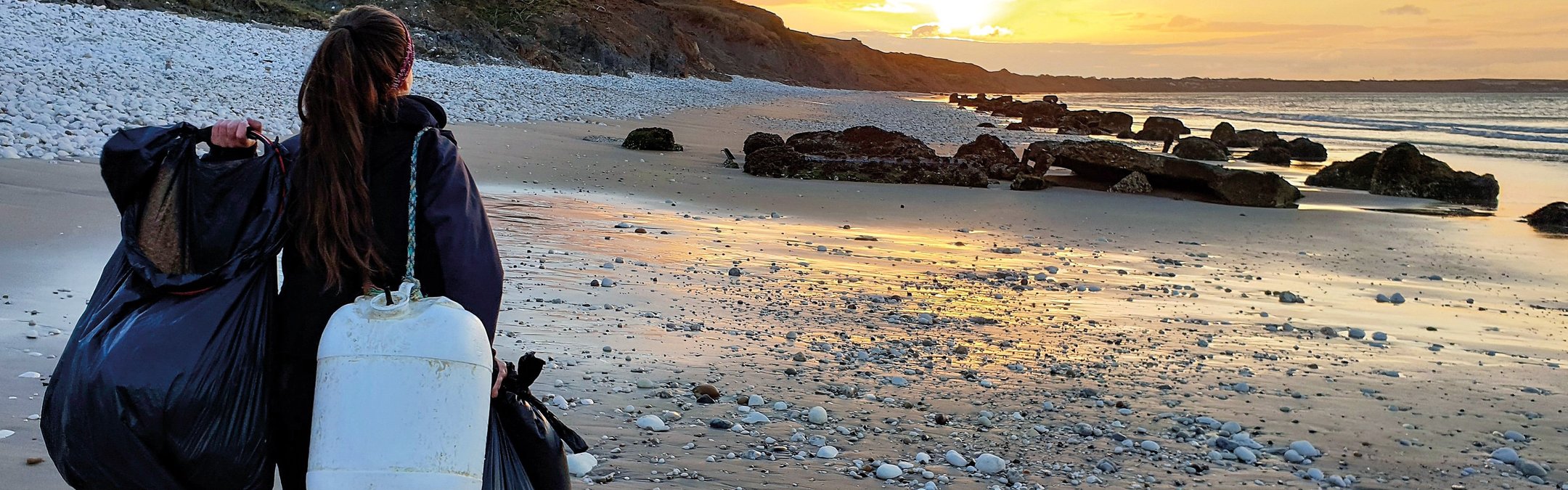 A girl walking away from the camera toward a setting sun. SHe is on a beach and is carrying a black bin bag and a white bucket.
