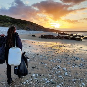 A girl walking away from the camera toward a setting sun. SHe is on a beach and is carrying a black bin bag and a white bucket.