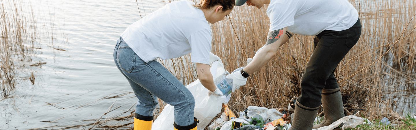 A man and woman putting litter into a white dustbin bag in front of a river. They ar wearing wellies and strong gloves.