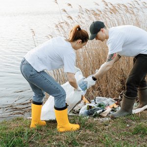 A man and woman putting litter into a white dustbin bag in front of a river. They ar wearing wellies and strong gloves.