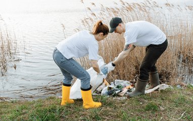 A man and woman putting litter into a white dustbin bag in front of a river. They ar wearing wellies and strong gloves.