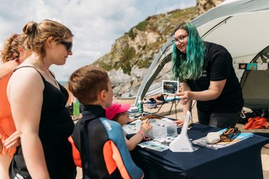 A volunteer at a Marine Conservation Society stall on a beach showing a picture of a shell to a woman and two children.