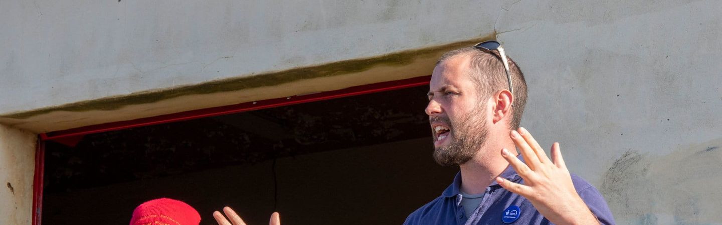 A man in a blue polo shirt, gestures with his hands while speaking to an audience. Another man in a red hat and a high-visibility vest stands next to him with his arms crossed.