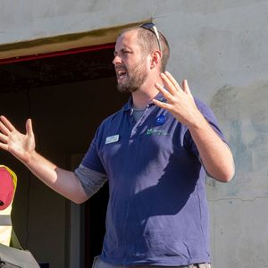 A man in a blue polo shirt, gestures with his hands while speaking to an audience. Another man in a red hat and a high-visibility vest stands next to him with his arms crossed.