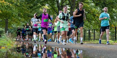 A group of runners participates in a race along a paved path lined with lush green trees. In the foreground, a large puddle reflects the vibrant colors of the runners' athletic gear.