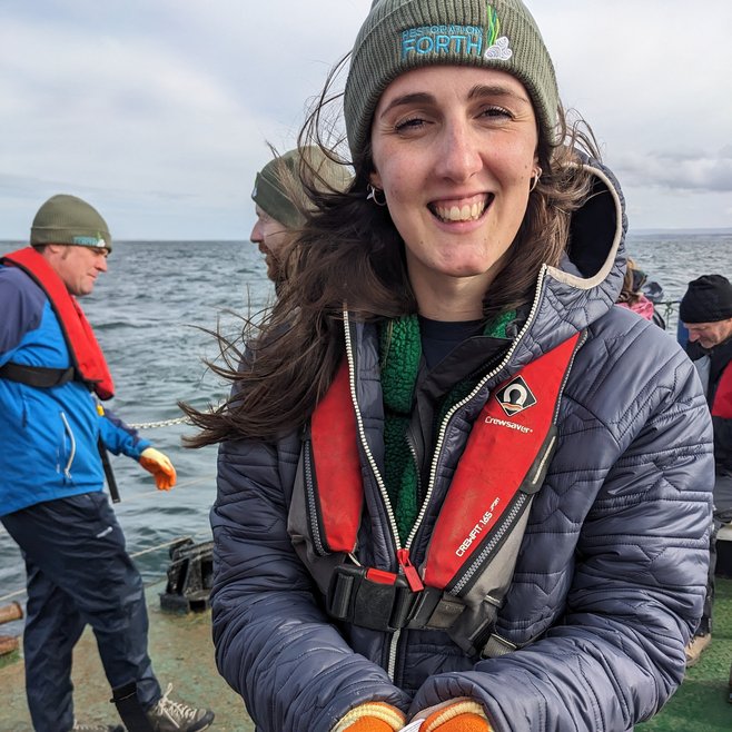 Caitlin Godfrey, Community Engagement Lead for Restoration Forth smiles at the camera from the shoreline with oysters in her hands.