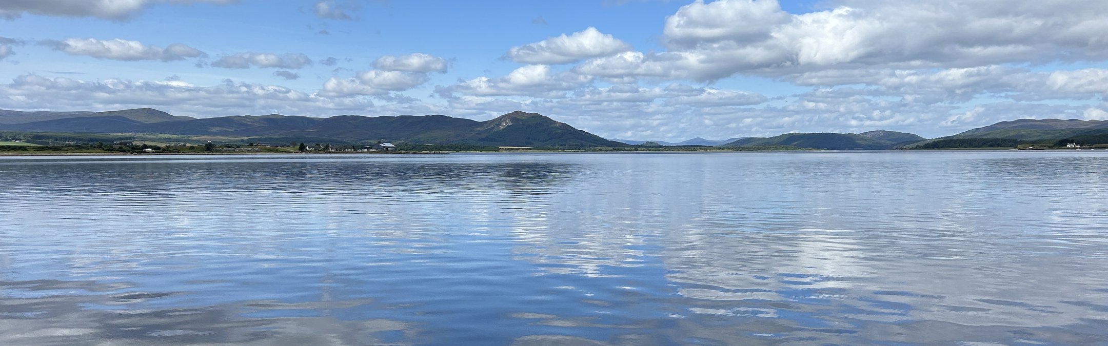 A photograph taken from the bank of the Dornoch Firth with far-reaching veiws across the calm water. The sky is blue with white clouds and across the water you can see the coastline where the Glenmorangie Distillery Company is based.