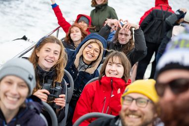 A group of smiling young people sitting in rows on a boat, one person in the foreground to the left is holding binoculars. The person in the background is making a heart with their fingers.