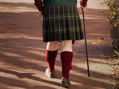 A person viewed from behind walking on a brick path, wearing a traditional Scottish kilt with a green, red, and yellow tartan pattern. They are also wearing knee-high burgundy kilt hose with tassels, brown leather shoes, and are using a dark walking cane.
