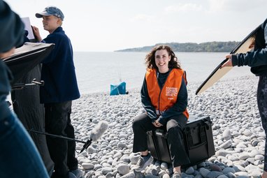 A Marine Conservation Society staff member in an orange vest sitting on a black box on a rocky beach, smiling at the camera, while a film crew works around them.