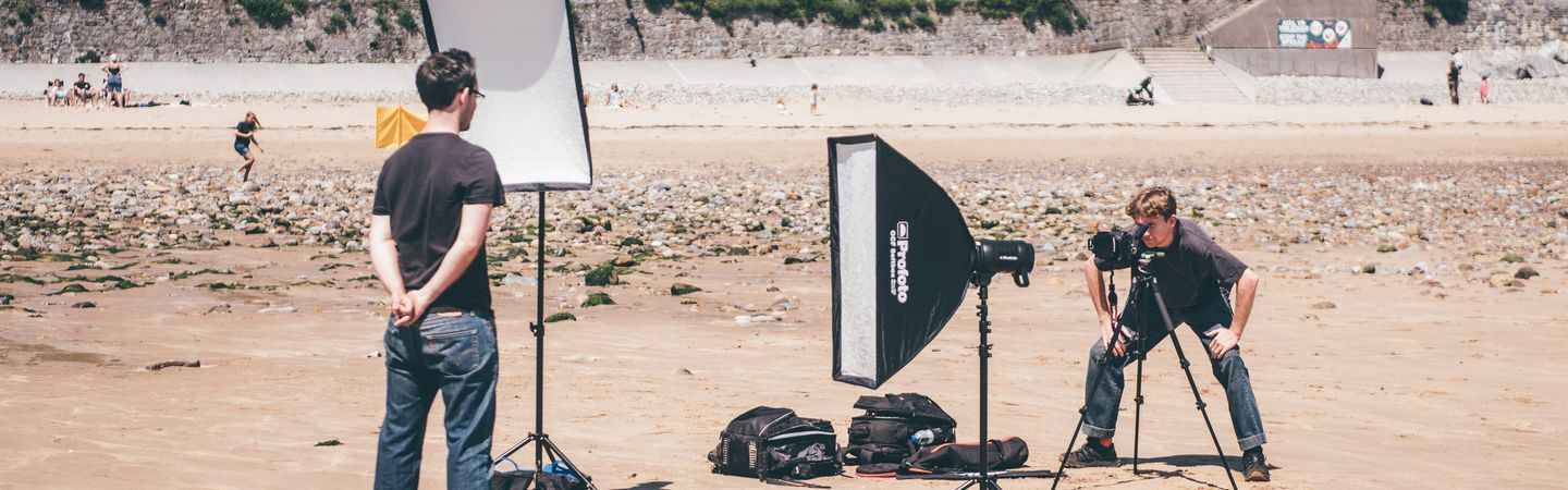 A man standin on a sandy beach looking at a second man who is crouching behind a camera on a tripod, with professional photography lighting equipment set up around them. In the background are a row of small, green-and-white striped beach huts.