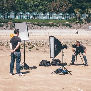 A man standin on a sandy beach looking at a second man who is crouching behind a camera on a tripod, with professional photography lighting equipment set up around them. In the background are a row of small, green-and-white striped beach huts.