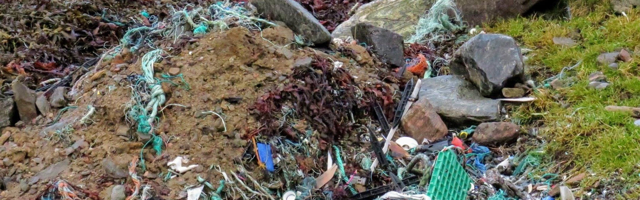 A close-up view of a beach covered in a large amount of marine litter, including tangled green and white plastic fishing ropes, various plastic debris, seaweed, soil, and rocks.