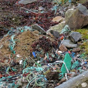 A close-up view of a beach covered in a large amount of marine litter, including tangled green and white plastic fishing ropes, various plastic debris, seaweed, soil, and rocks.