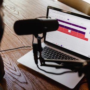 A female sits at a desk. On the desk is a laptop, and next to the laptop is a podcast microphone.
