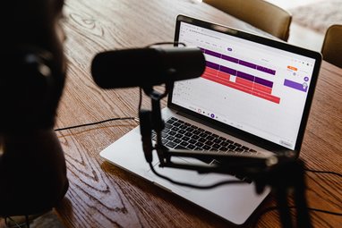 A female sits at a desk. On the desk is a laptop, and next to the laptop is a podcast microphone.