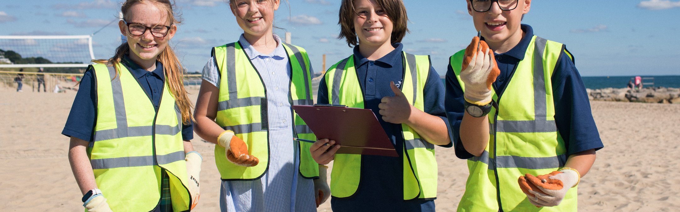 4 school children looking at the camera wearing hi-vis jackets and gloves on. The girl on the far left is holding a litter picker and the boy that is centre right is holding a clipboard. They are on a beach