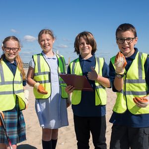 4 school children looking at the camera wearing hi-vis jackets and gloves on. The girl on the far left is holding a litter picker and the boy that is centre right is holding a clipboard. They are on a beach