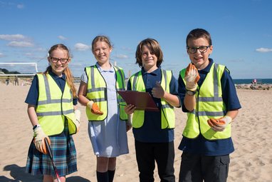 4 school children looking at the camera wearing hi-vis jackets and gloves on. The girl on the far left is holding a litter picker and the boy that is centre right is holding a clipboard. They are on a beach