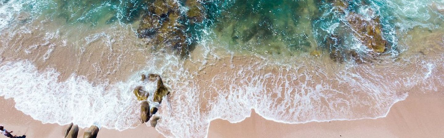 Shown from above, sea waves are spilling over sand and rocks. The sea is a deep green while the foamy surf is breaking.