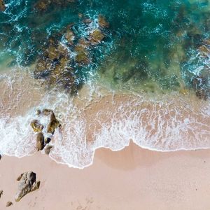 Shown from above, sea waves are spilling over sand and rocks. The sea is a deep green while the foamy surf is breaking.
