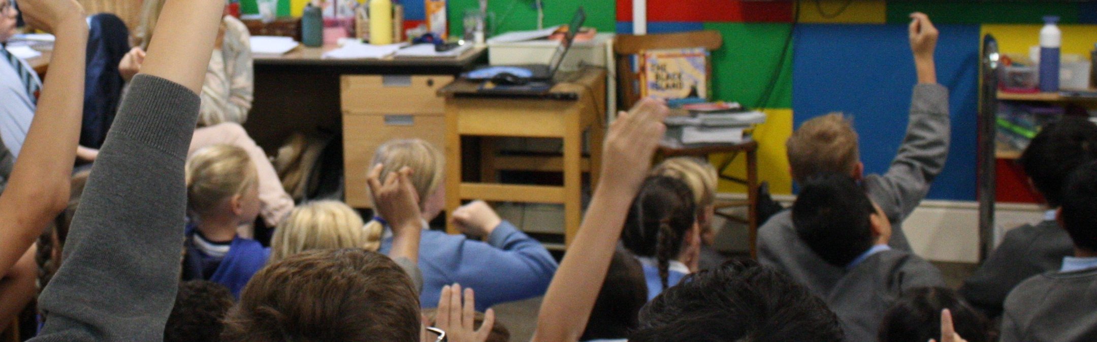 A classroom of children sitting on the floor looking at a screen, some have their hands up