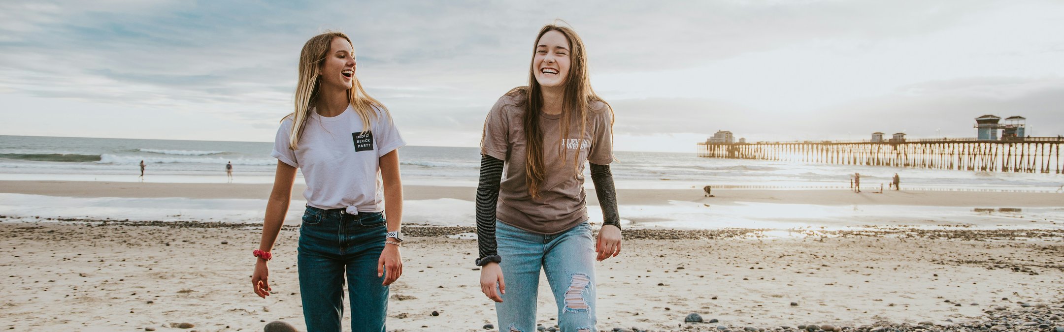 2 teenage girls laughing on the beach. It is a rocky beach with a small pier in the distance and appears to be dusk.
