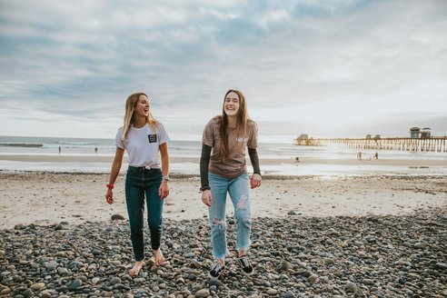 2 teenage girls laughing on the beach. It is a rocky beach with a small pier in the distance and appears to be dusk.