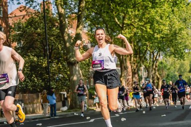 A young woman with hair in a pony tail celebrates with a joyful shout and raised arms while running the Hackney Half