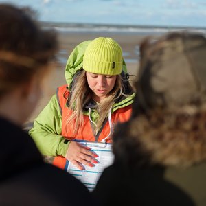 A woman wearing a lime green beanie and orange safety vest is looking down at a sheet on a clipboard. She is on a beach and is explaining the sheet to 2 people in the foreground