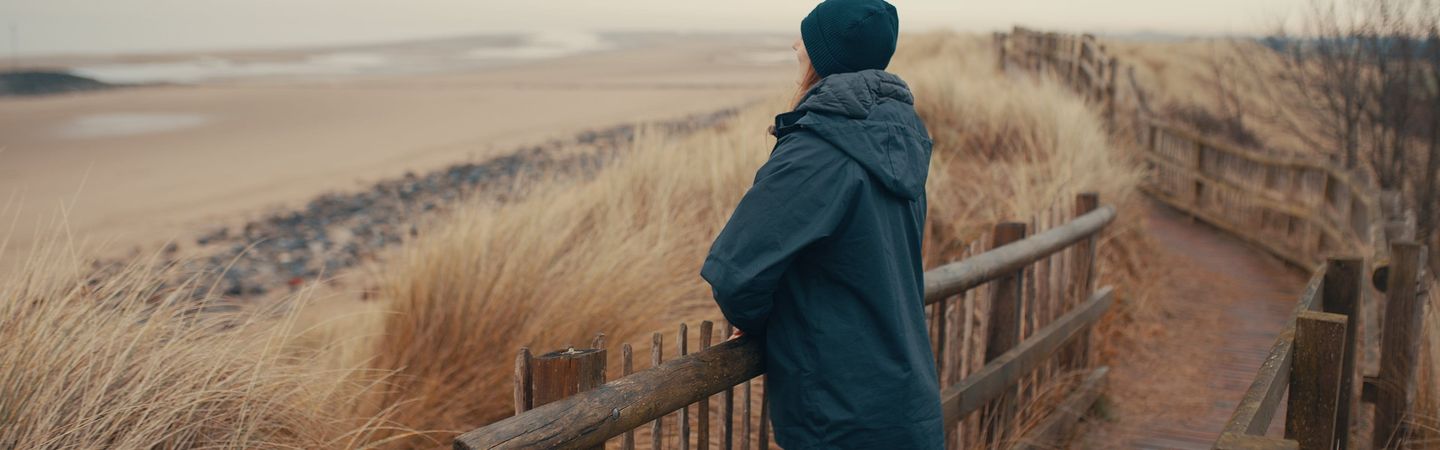 Woman standing over a fence looking towards a beach. She is wearing a dark coat and beanie. She is facing away from the photographer.