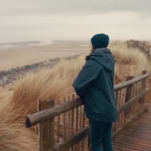 Woman standing over a fence looking towards a beach. She is wearing a dark coat and beanie. She is facing away from the photographer.
