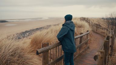 Woman standing over a fence looking towards a beach. She is wearing a dark coat and beanie. She is facing away from the photographer.