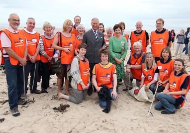King Charles III attends a Bamburgh beach clean in 2012. He is smiling in the middle of the photo with around 20 volunteers in orange vests. Young volunteers are kneeling and sat down in the foreground.