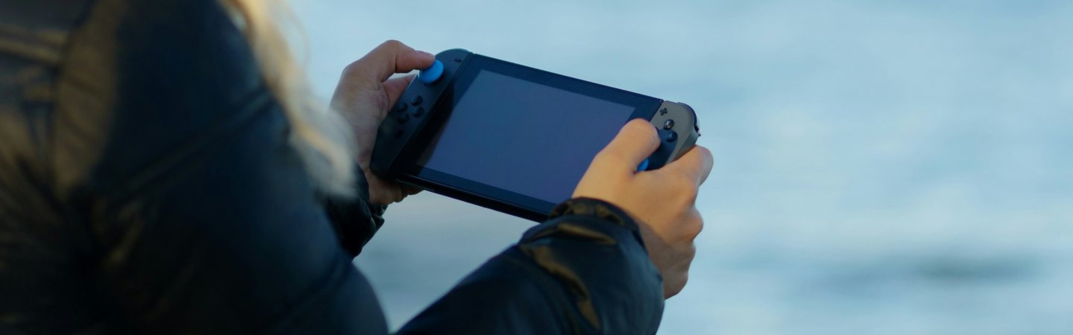An over the shoulder shot of a woman with blonde hair holding a Nintendo switch. In the background appears to be a lake or ocean.