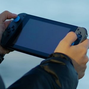 An over the shoulder shot of a woman with blonde hair holding a Nintendo switch. In the background appears to be a lake or ocean.