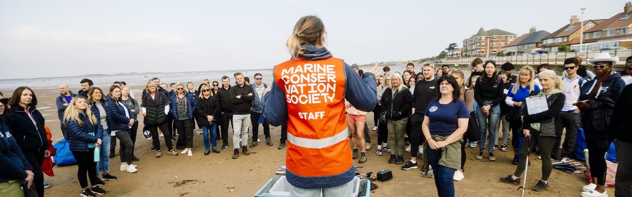 A woman wearing a hi-vis orange vest has her back to the camera. She is in front of a group of volunteers. Her vest reads Marine Conservation Society.