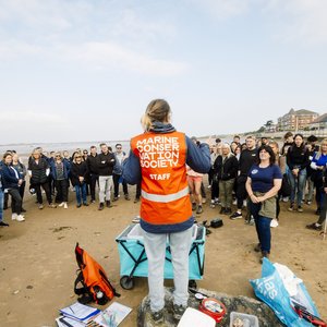 A woman wearing a hi-vis orange vest has her back to the camera. She is in front of a group of volunteers. Her vest reads Marine Conservation Society.