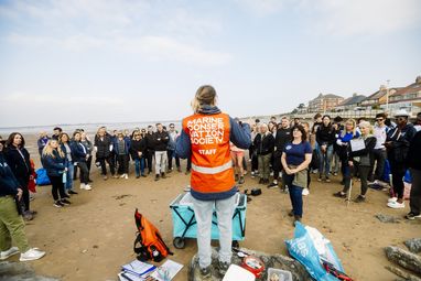 A woman wearing a hi-vis orange vest has her back to the camera. She is in front of a group of volunteers. Her vest reads Marine Conservation Society.