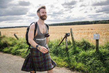 A bearded man with a handlebar mustache smiles as he walks along a rural path. He is wearing a grey tank top, a traditional tartan kilt, and a black backpack. The background features a golden field under a cloudy sky.