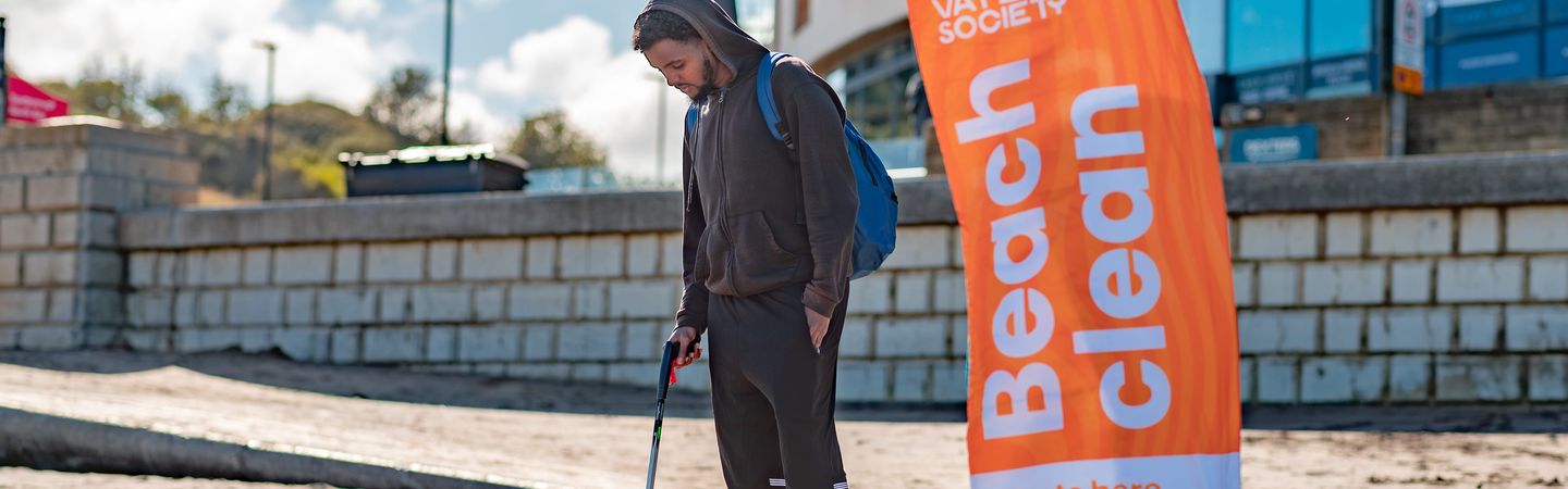 A male using a litter picker to pick up a plastic bottle on a beach. A Marine Conservation Society banner is to the right of the man.