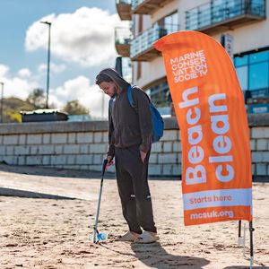 A male using a litter picker to pick up a plastic bottle on a beach. A Marine Conservation Society banner is to the right of the man.