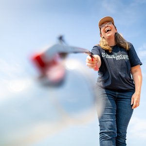 A person wearing a navy blue Marine Conservation Society t-shirt, jeans, and a tan cap laughing while using a litter picker. The camera angle is low, looking up at the person against a bright, clear blue sky. A blurred plastic bottle is in the foreground.