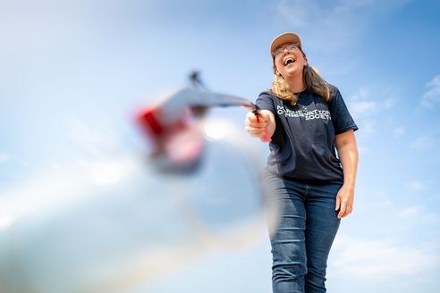 A person wearing a navy blue Marine Conservation Society t-shirt, jeans, and a tan cap laughing while using a litter picker. The camera angle is low, looking up at the person against a bright, clear blue sky. A blurred plastic bottle is in the foreground.