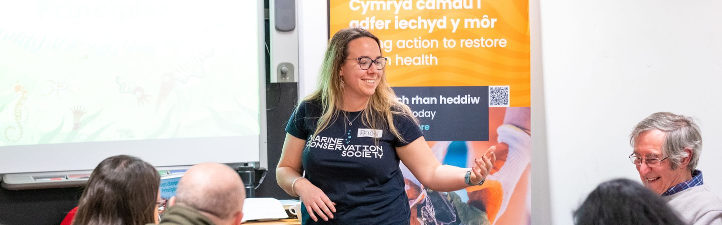 A woman wearing a Marine Conservation Society t-shirt speaking to an adult audience in front of a banner that reads 'Cymryd camau i adfer iechyd y môr' and 'taking action to restore marine health'."