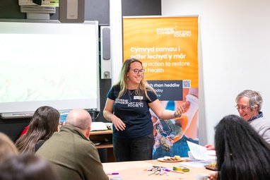 A woman wearing a Marine Conservation Society t-shirt speaking to an adult audience in front of a banner that reads 'Cymryd camau i adfer iechyd y môr' and 'taking action to restore marine health'."