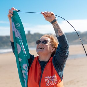 A woman wearing snglasses and an orange hi-vis vest is placing a teal flag onto a pole. Behind he is a beach.