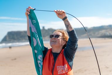 A woman wearing snglasses and an orange hi-vis vest is placing a teal flag onto a pole. Behind he is a beach.
