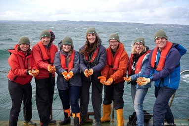 Seven scientists and Marine Conservation Society staff involved in the Restoration Forth project stand smiling on the Firth of Forth shore. They are all wearing protective orange gloves with European oysters in their spread-out palms.